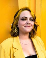 Megan Joy is standing in front of a yellow background wearing a yellow blazer and smiling